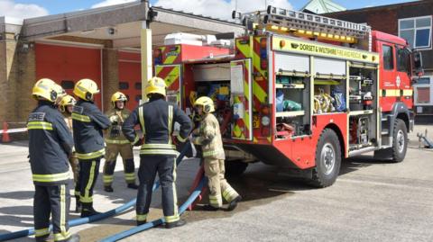 A crew of five firefighters in uniform standing outside a fire engine. The vehicle is parked at a fire station. There are two blue hosepipes coming from the back of the fire engine.