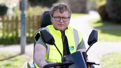 A man with short, brown hair and glasses wearing a yellow high-vis coat. He is sitting on a mobility scooter.