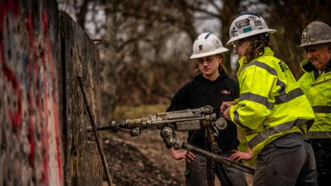 Three people dressed in a protective clothing with plastic helmets using a drill to drill into a wall. One person is using the drill while the other two are observing. They appear to be in a rural location with trees in the background.  