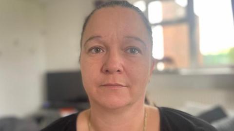 Head-and-shoulders image of a Louise Butcher looking directly at the camera in a softly lit room, with a blurred television and window in the background.