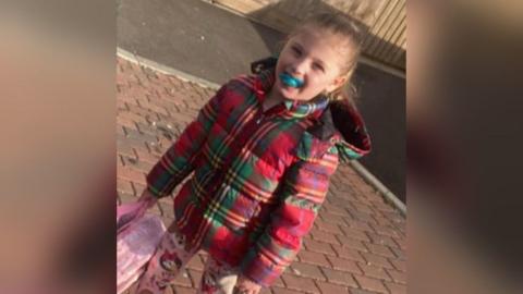 A little girl with blond hair. She has a red checked puffer jacket on and a dummy in her mouth. She is holding a pink backpack. 