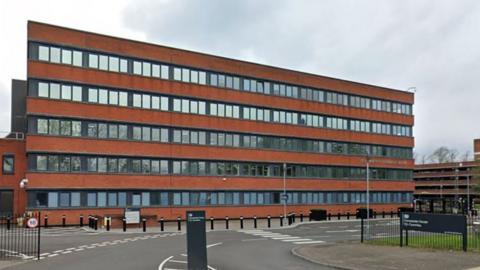 The Companies House HQ in Cardiff. It is a five-storey red brick building with long rows of windows on each level. There is a multi-storey car park to the right and black gates / fence on both sides of the road into the complex.