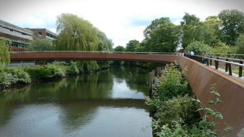 Walkers crossing Jarrold Bridge in Norwich. The bridge is a rusty colour and trees line both sides of the river.