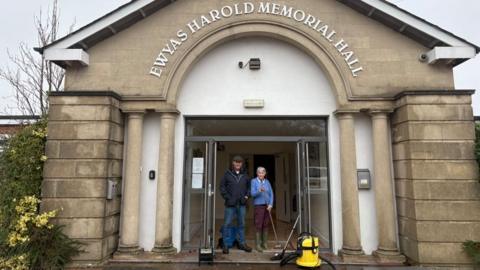 A man and a woman standing outside a memorial hall. The hall is made from stone and has two columns on either side of the entrance. The man and woman are cleaning up after flooding so they are wearing wellies and there is a broom as well as mop bucket and hoover.