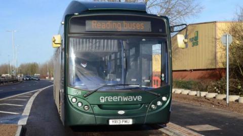 Green bus travelling towards the camera in a bus lane.  A road and traffic can be seen behind.