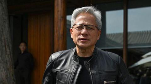 Jensen Huang wearing a black leather jacket and glasses, standing outside a wood and glass building