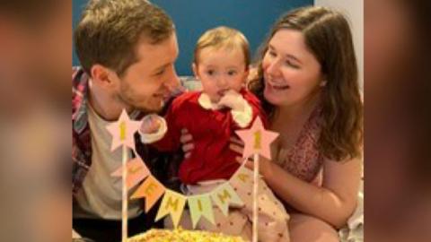 A man, a woman and a baby sit behind a cake with a candle in it