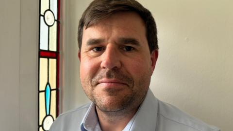 Ian Hewitt with brown eyes, dark brown stubble and brown hair standing in front of a door with a red and blue stained glass feature 