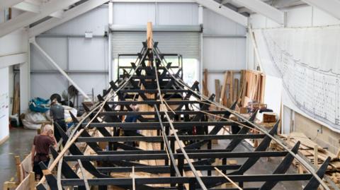 The skeleton of a ship made of wooden panels is pictured inside a large warehouse. People can be seen working on the ship while wooden panels rest all around it.