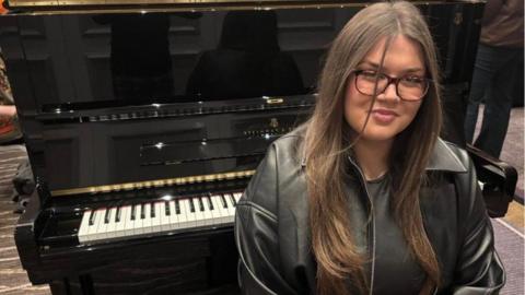 Grace Matthews with long brown hair and glasses sits in front of an open Steinway piano. 