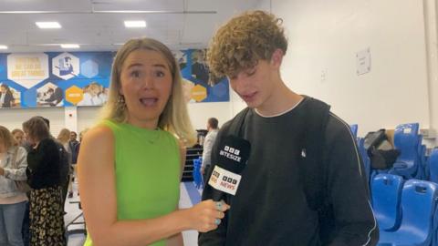 A ˿ reporter wearing a green top grins shocked as a cardiff secondary student opens his GCSE results