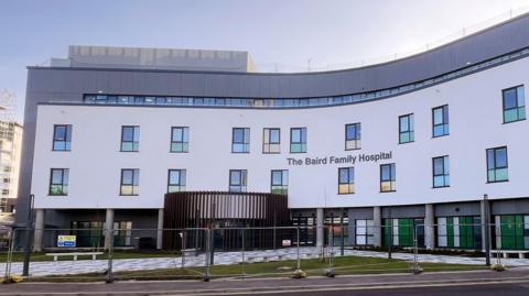 The hospital is a large, four storey white and grey building, part of the building is blue and extends from the front of the hospital. 