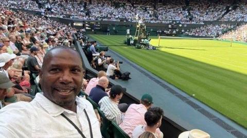A man in a white collared shirt with a black lanyard around his neck is taking a selfie. He is sitting inside a tennis court, with a rows of audiences in the background.