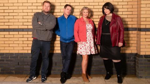 Four members of the cast of Gavin and Stacey reunite for an official cast photo: Smithy, Gavin, Stacey and Nessa. They are stood against a brick wall. Smithy (played by James Corden) is wearing a grey jacket and blue jeans and has his arms crossed, Gavin (played by Matthew Horne) is next to him, wearing a blue jacket with his hands in the pockets and blue jeans. Next to him is Stacey (played by Joanna Page), she is wearing brown cowboy boots, a white floral dress and a pink jacket, and she is stood next to Nessa (played by Ruth Jones), who is wearing a burgundy leather jacket and a black dress with long black boots. 