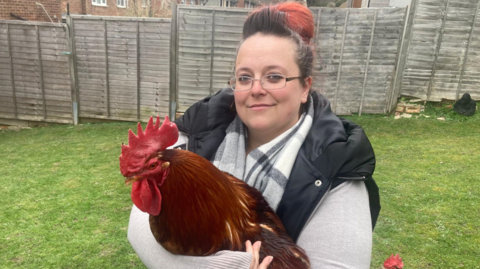 Claire Tarr is holding a large cockerel in her garden. It is russet brown with a red face. She is smiling and is wearing a grey and white chequered scarf around her neck.