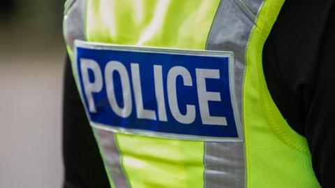 The back of a police officer, showing their yellow high-vis vest with the word 'police' in capital letters across the back. The picture is zoomed into this area, with the background out of focus. 