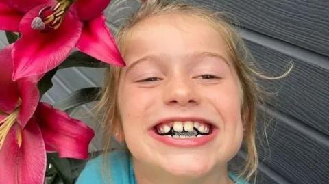 Scarlett Rossborough, a young girl, smiles at the camera while standing next to a large pink flower