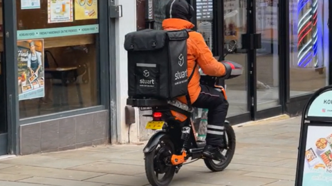 Man in orange jacket rides electric bike through city centre with large black food delivery rucksack on his back