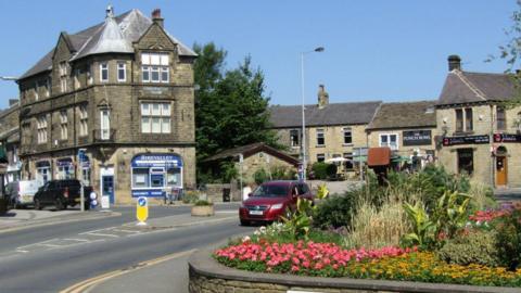 The centre of Silsden in Bradford in West Yorkshire, with a large stone building on the left, The Punch Bowl pub on the right and a maroon car driving on the road between the two. Several stone buildings are in the background and a flowerbed with a low wall with the sign 'Wesley Place' is in the foreground.