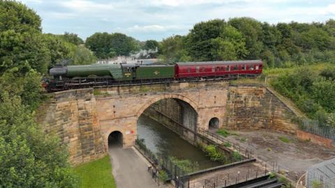 The green Flying Scotsman steam train pulling a red carriage on a track over a stone archway bridge over a canal. Thick trees line either side. 