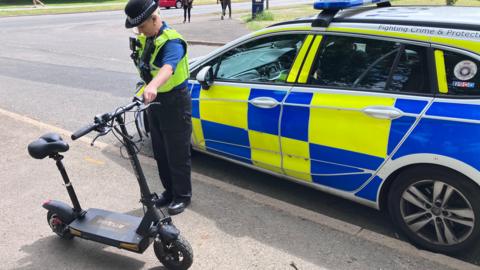 A female police officer stands on the pavement next to a police car, holding a black e-scooter. She is wearing a black uniform and a black police hat.