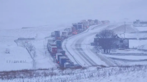 Traffic queues in snowy conditions on a blocked section the A66 in Durham, North East England, near to the border with Cumbria. There is heavy snowfall and a thick line of traffic.