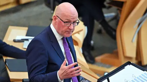 a bald man with glasses is wearing a blue suit, white shirt and purple tie as he answers a questions in the parliament chamber