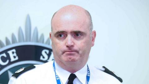 A policeman wearing a white shirt with a police badge on the right side pocket. A name tag on the left side and a black tie. A blue lanyard reading staff is also around his neck.