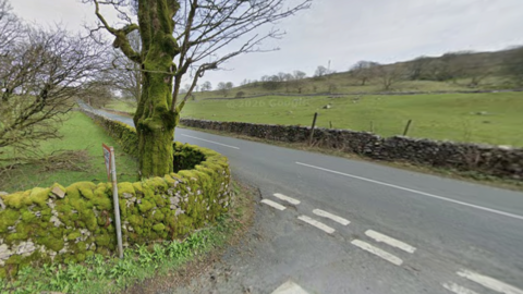 A quiet country road running through open farmland under a grey, overcast sky. A moss-covered stone wall and a large tree stand close to the roadside, with grassy fields stretching out beyond. White road markings are visible at a small junction in the foreground. 