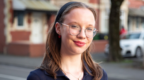 A girl with brown hair and glasses, wearing a blue top, smiles at the camera