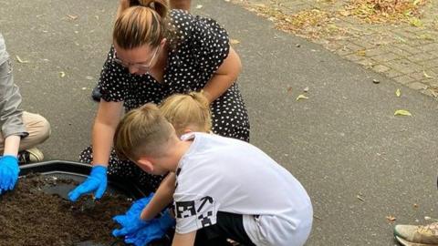 A woman and two children are sat on the floor gardening. They have blue gloves on and are putting their hands in some soil. 