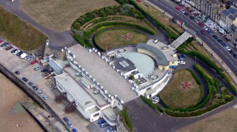 An aerial shot of a circular building surrounded by gardens and bordered by a road and a beach.