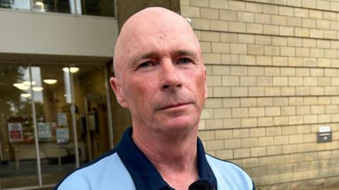 A photo of Northamptonshire councillor Raymond Connolly. He is standing outside Northampton Crown Court and is facing the camera without an expression on his face. He is wearing a blue top with a navy collar.