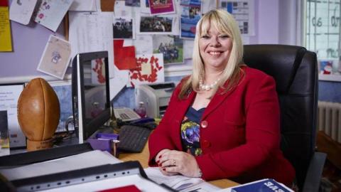 Joy Ballard a blonde haired woman, sat at her desk and smiling at the camera, wearing a red jacket and dress.