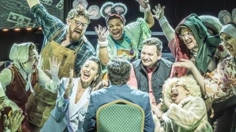 A scene from the musical Bank of Dave. A man in a suit is seated on a chair with his back to the camera while characters wearing different costumes representing different characters sing in front of him.