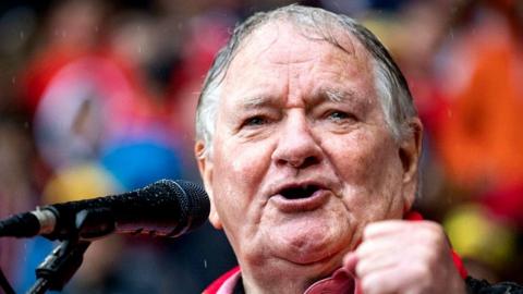 Close up of Dafydd Iwan's face as he sings into a microphone. He has tears in his eyes and is holding an out-of-focus fist slightly in front and to the side of his face. Behind are football blurry fans as this is taken in Cardiff City Stadium