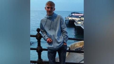 A man wearing a blue zip up hoody is stood with an elbow leaning on a railing with the sea and a pier behind him.