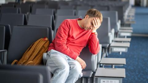 A man in a red jumper sits with his head in his hand at a bank of airport teminal seats.