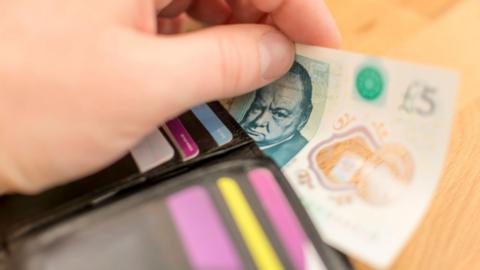 A close-up image of a hand removing a five pound note from a black wallet. Generic purple and yellow cards can be seen.