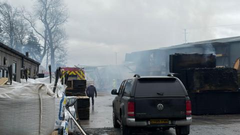 Plumes of white smoke fill the sky. On the right side of the image is tin shed and beside of there are a number of pallets of wood. A fire engine and an officer in a yellow suit is tackling the blaze. A black car is parked up in the yard. 