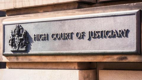 A rectangle-shaped grey sign on an exterior, stone block wall of the High Court in Edinburgh. The sign has a royal crest and the words: "High Court of Justiciary".