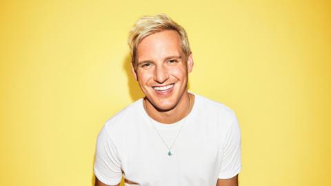 Jamie Laing smiles in front of a yellow background - he has short blond hair and wears a white T-shirt 