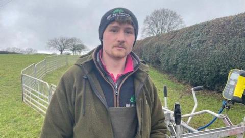 Ollie Raybould is wearing a woolly black hat with his curly brown fringe hanging out of it. He is wearing a green fleece jacket and pink t-shirt underneath it