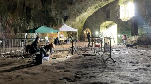 Archaeologists digging at an excavation in a large cave, with ancient walls visible in the background