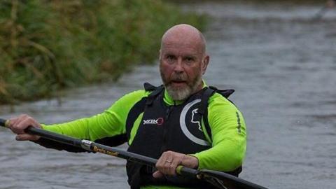 Neil Armstrong Boast kayaks on a river. He is bald and wears a bright green top with a life jacket on. He holds the paddle in both hands.