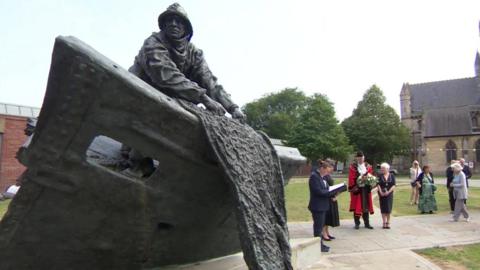 People gather as a woman reads a tribute next to a statue of a fisherman pulling nets over the side of a boat.