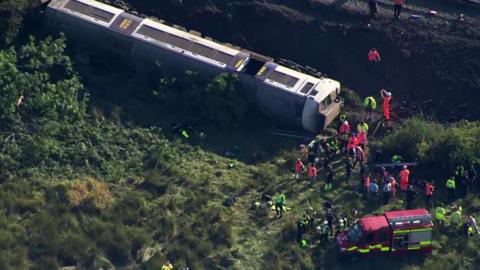 an aerial view of a train carriage on it's side alongside railway tracks. Multiple emergency workers and vehicles are surrounding the carriage. 