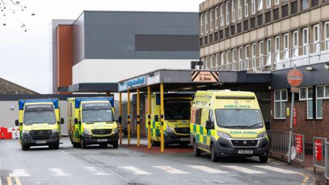Three NEAS ambulances parked outside Darlington Memorial Hospital. It is a two-storey building with windows. A more modern grey building is visible in the background.
