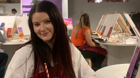 A woman with long dark red hair smiles while sitting at a table. Behind her people can be seen sat on white tables with easels propped up on them while painting. 