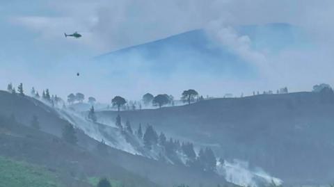 A helicopter flies above Cairngorms national park with trees covered by smoke
